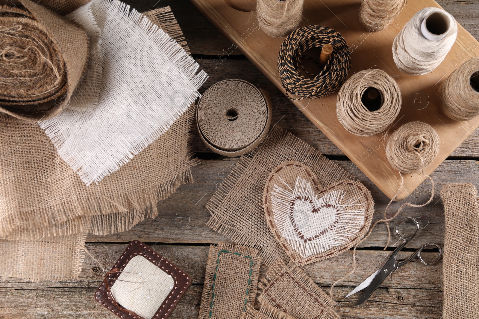 Photo of Pieces of burlap fabric, spools of twine and different sewing tools on wooden table, flat lay