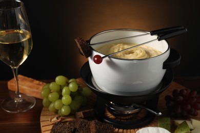 Forks with pieces of grape, bread, fondue pot with melted cheese, wine and snacks on table, closeup