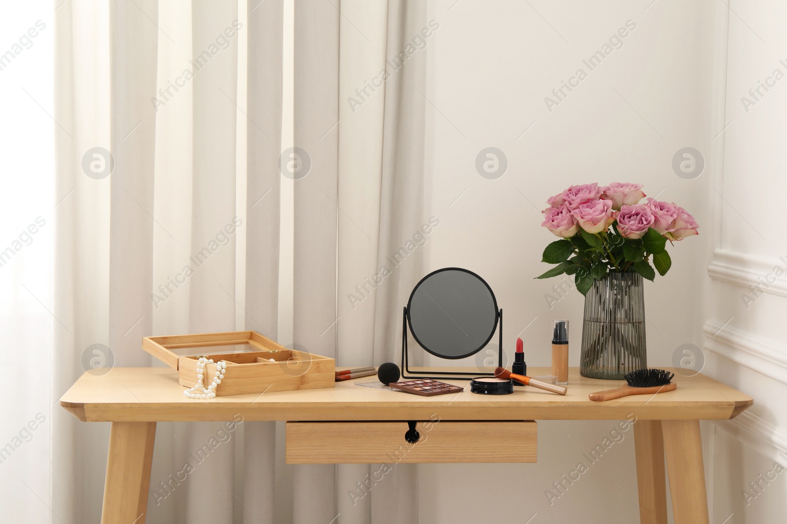 Photo of Mirror, cosmetic products, box of jewelry and vase with pink roses on wooden dressing table in makeup room