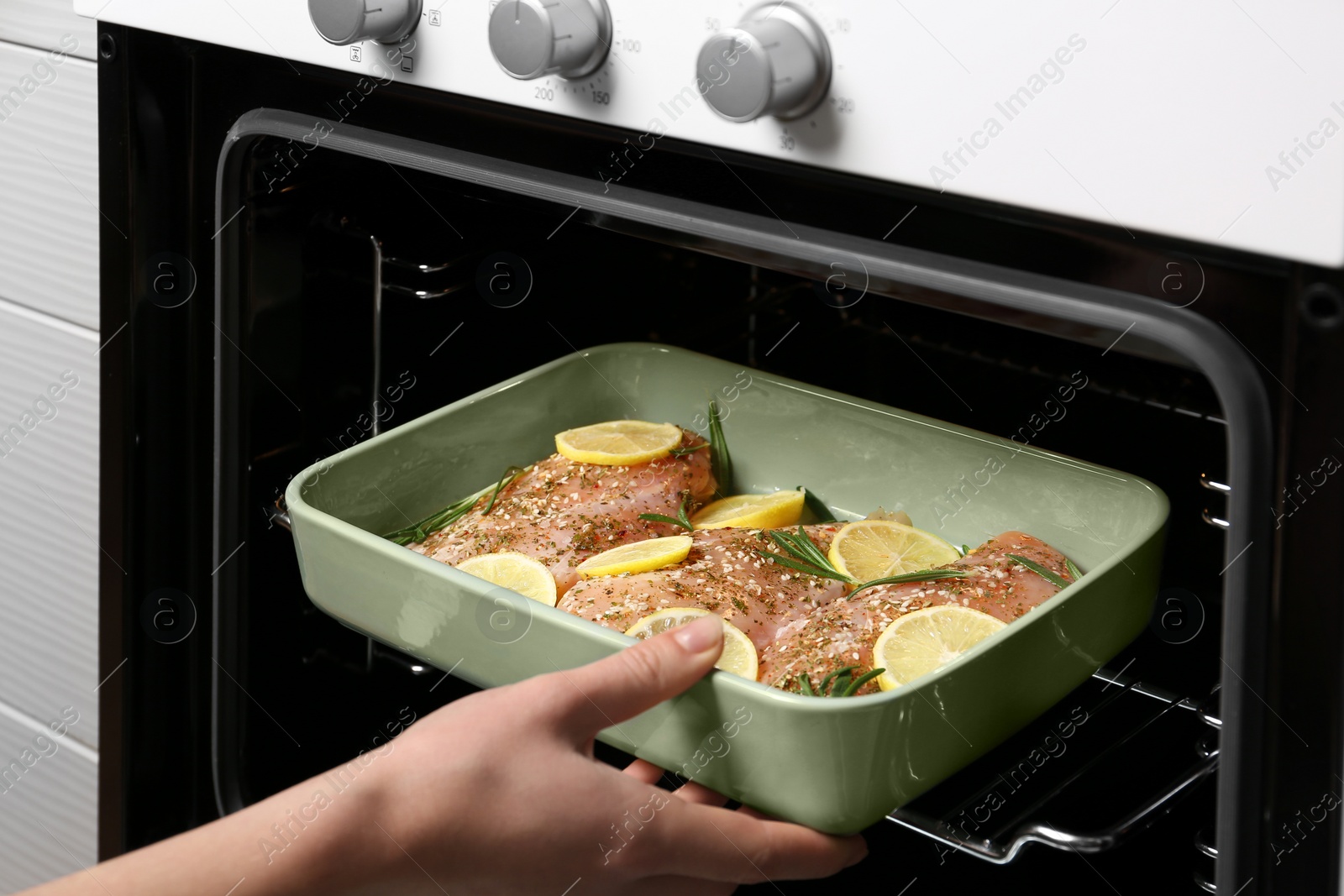 Photo of Woman putting chicken breasts with lemon and rosemary into oven, closeup