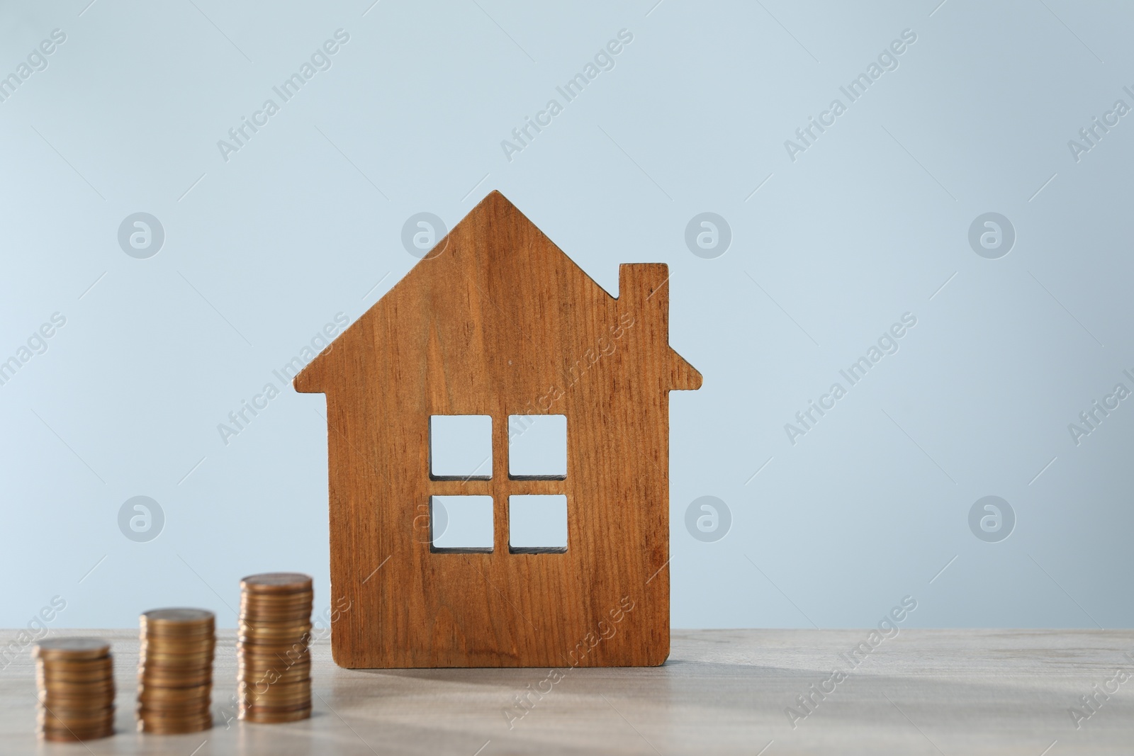 Photo of House model and stacked coins on wooden table against light blue background, space for text