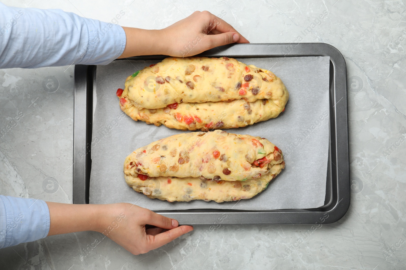 Photo of Woman holding baking tray with raw homemade Stollens at grey table, closeup. Traditional German Christmas bread