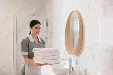 Photo of Young chambermaid holding stack of fresh towels in bathroom