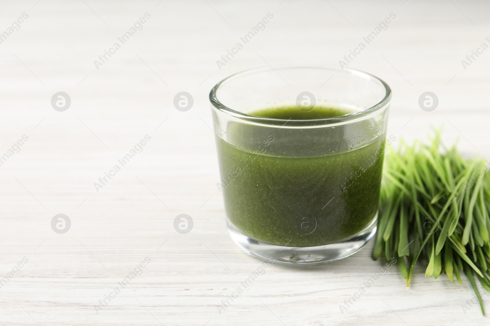 Photo of Wheat grass drink in glass and fresh sprouts on white wooden table, closeup. Space for text