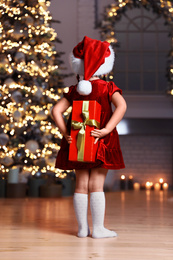 Cute little child wearing Santa hat hiding Christmas gift behind her back at home