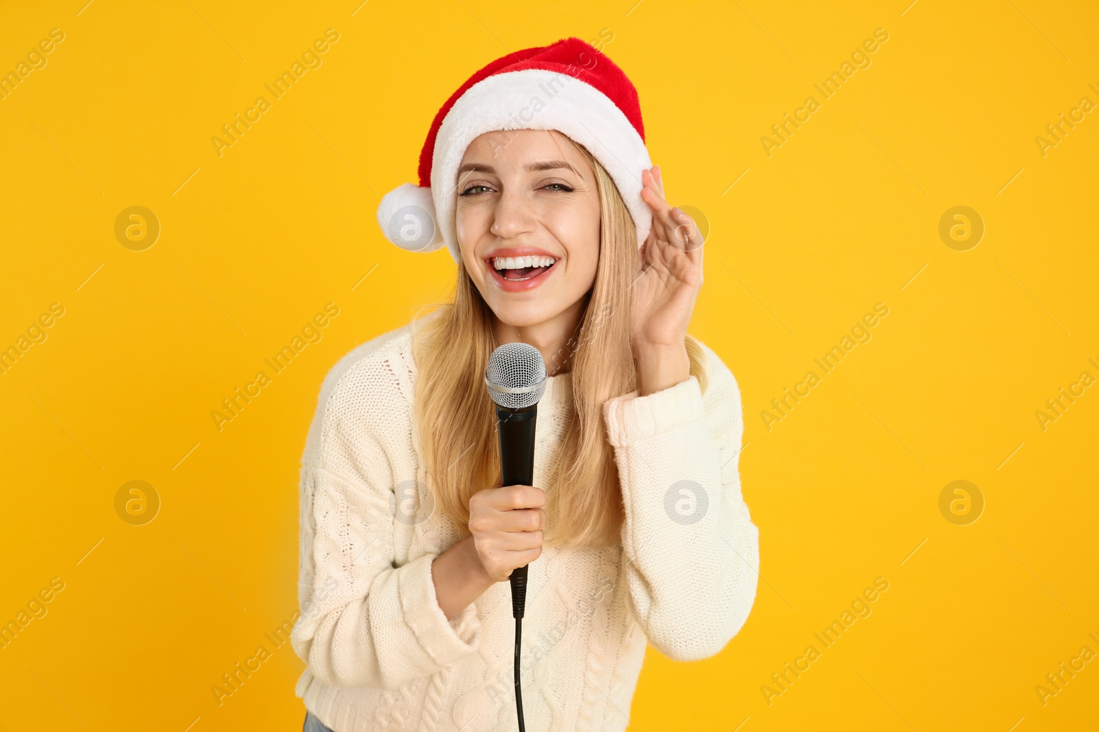 Photo of Emotional woman in Santa Claus hat singing with microphone on yellow background. Christmas music