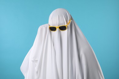 Photo of Stylish ghost. Person covered with white sheet in sunglasses on light blue background