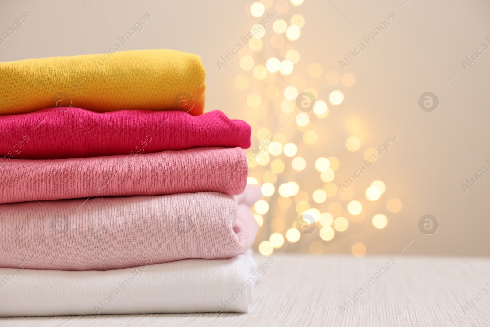 Stack of folded clothes on wooden table against beige background with blurred lights, space for text Photo of Stack of folded clothes on wooden table against beige background with blurred lights, space for text