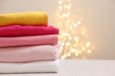 Photo of Stack of folded clothes on wooden table against beige background with blurred lights, space for text
