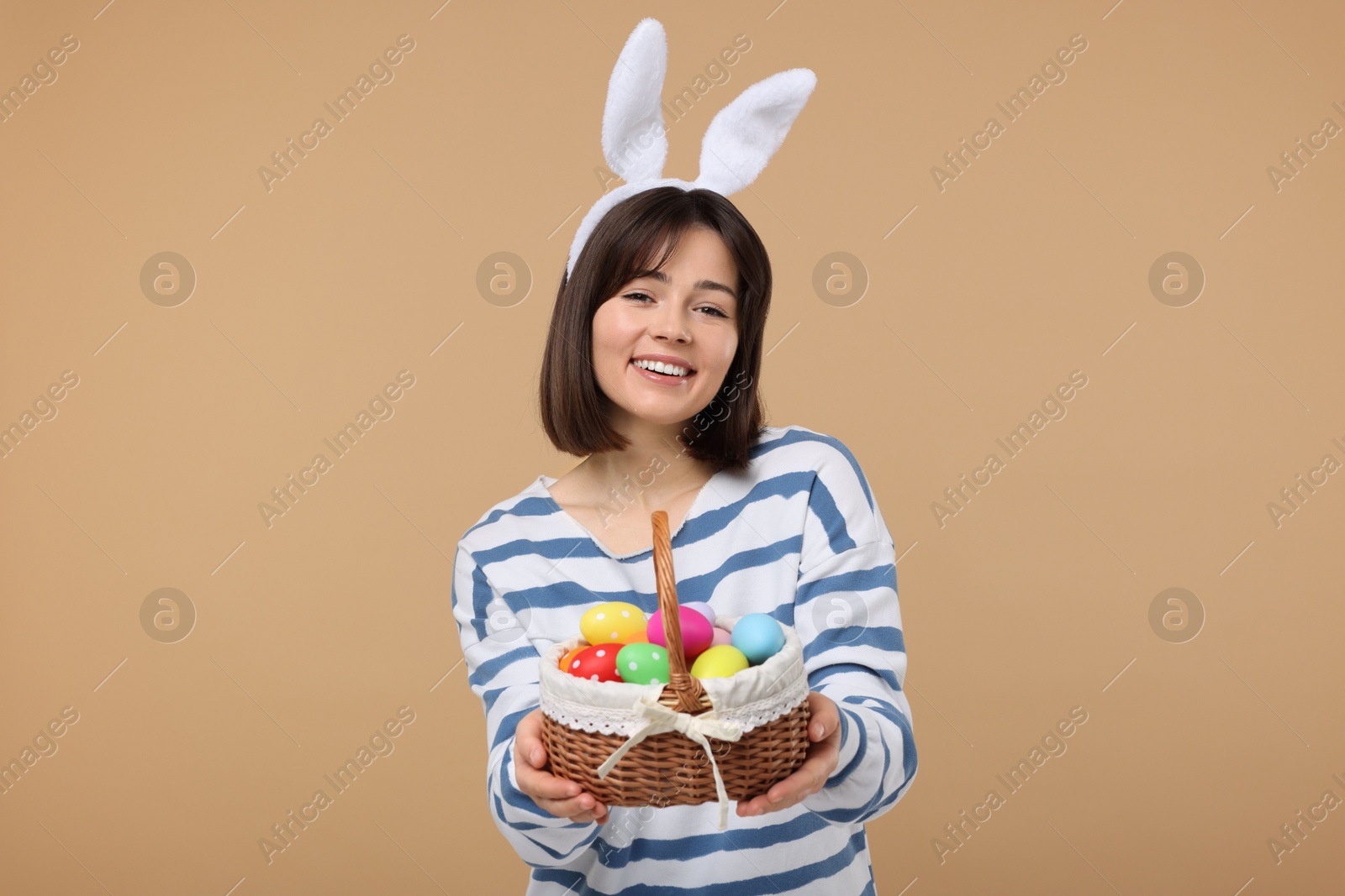 Photo of Easter celebration. Happy woman with bunny ears and wicker basket full of painted eggs on beige background