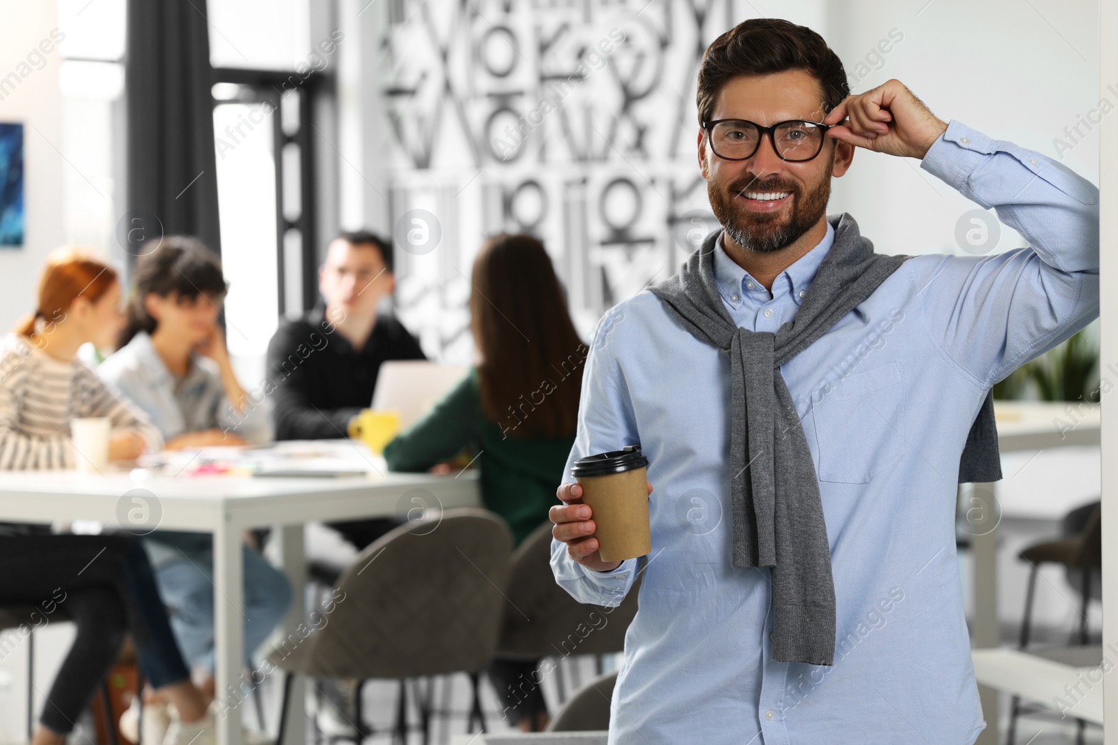 Photo of Team of employees working together in office. Happy man with paper cup of drink indoors, space for text