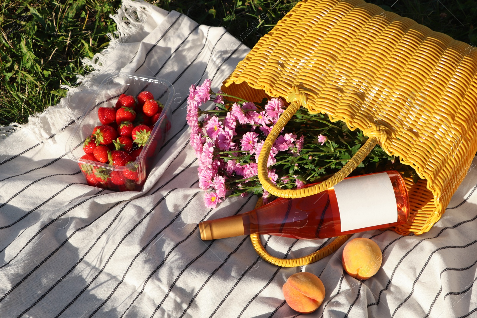 Photo of Yellow wicker bag with beautiful flowers, bottle of wine and food on picnic blanket outdoors