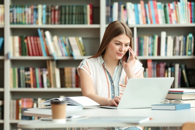 Young woman talking on phone and working with laptop at table in library. Space for text