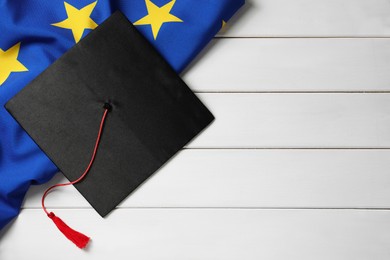 Photo of Black graduation cap and flag of European Union on white wooden table, flat lay. Space for text