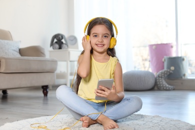 Photo of Cute child with headphones and mobile phone on floor indoors