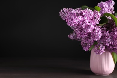 Photo of Beautiful lilac flowers in vase on wooden table against black background. Space for text