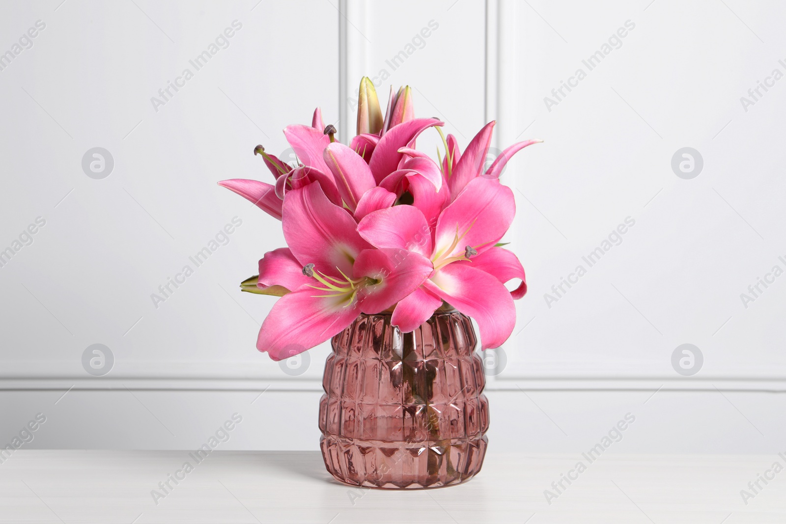 Photo of Beautiful pink lily flowers in vase on wooden table against white wall
