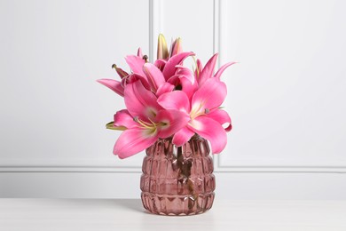 Photo of Beautiful pink lily flowers in vase on wooden table against white wall