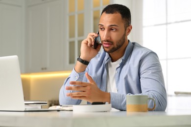 Photo of Young man talking on smartphone while working with laptop at desk in kitchen. Home office