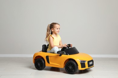 Photo of Cute little girl driving children's electric toy car near grey wall indoors