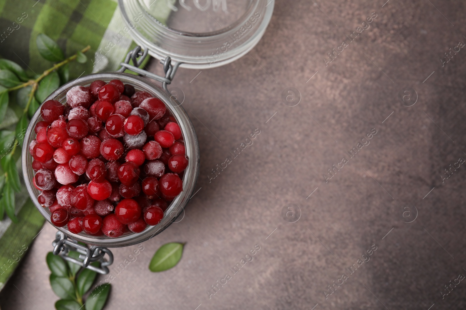 Photo of Frozen red cranberries in glass jar and green leaves on brown textured table, top view. Space for text