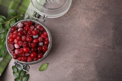 Photo of Frozen red cranberries in glass jar and green leaves on brown textured table, top view. Space for text