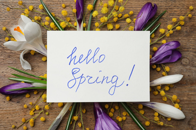 Photo of Card with words HELLO SPRING and fresh flowers on wooden table, flat lay