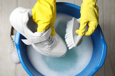 Photo of Woman with gloves and brush cleaning stylish sneakers in wash basin, top view