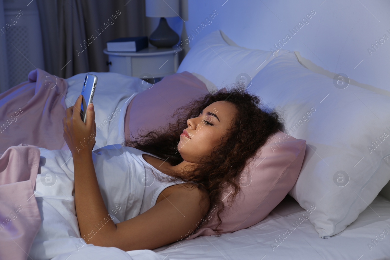Photo of Young African-American woman with mobile phone at night. Bedtime