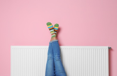 Photo of Woman warming legs on heating radiator near color wall
