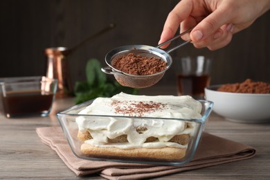 Photo of Woman pouring powdered cocoa onto tiramisu cake at wooden table, closeup
