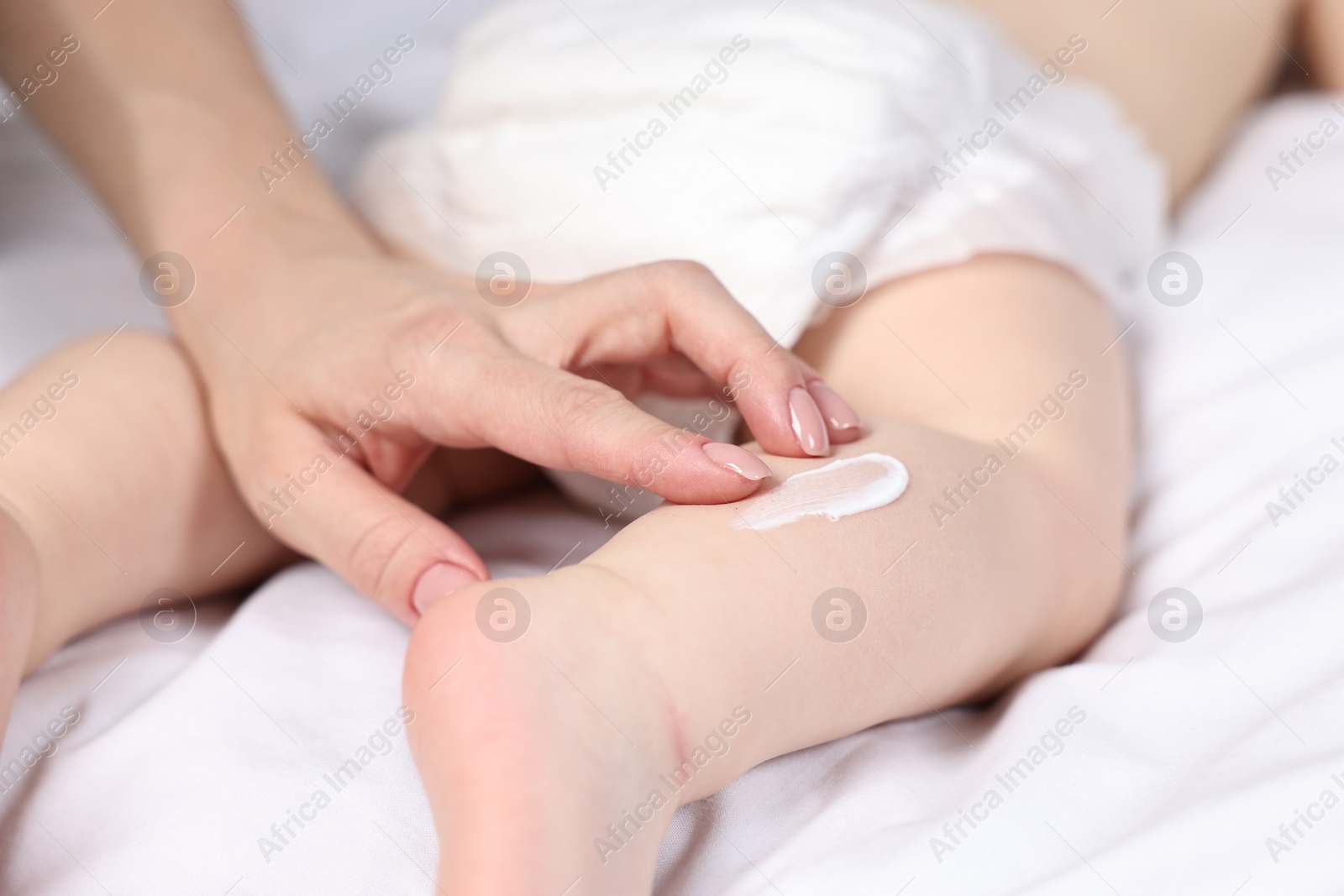 Photo of Woman applying body cream onto baby`s leg on bed, closeup