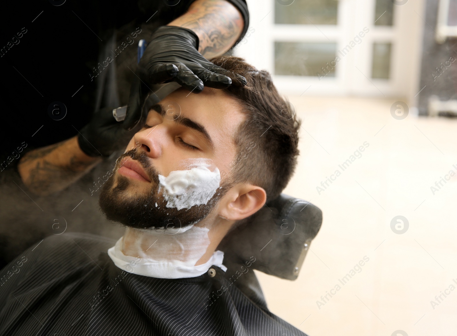 Photo of Professional hairdresser shaving client with straight razor in barbershop