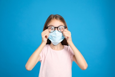 Photo of Little girl wiping foggy glasses caused by wearing disposable mask on blue background. Protective measure during coronavirus pandemic