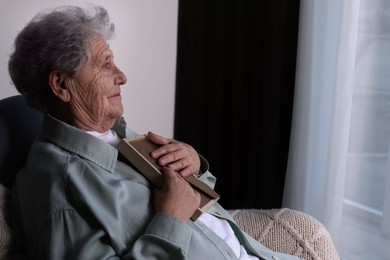 Photo of Elderly woman with photo frame in armchair indoors, space for text. Loneliness concept