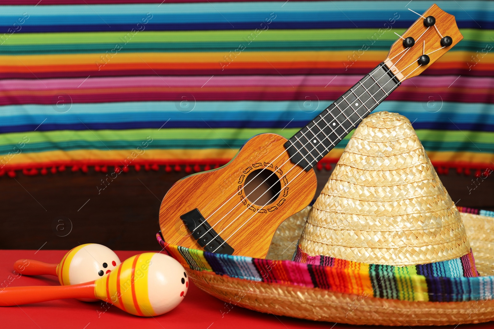 Photo of Mexican sombrero hat, ukulele and maracas on red table, closeup
