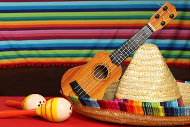 Photo of Mexican sombrero hat, ukulele and maracas on red table, closeup