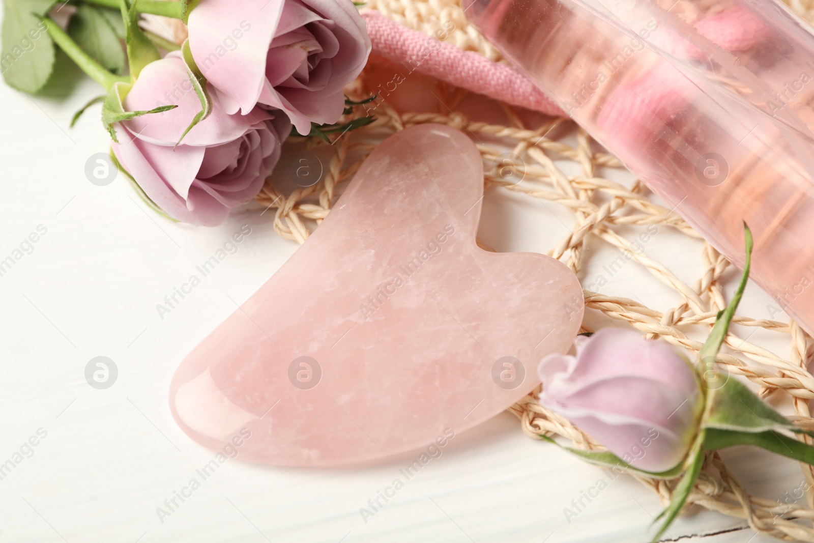 Photo of Rose quartz gua sha tool, flowers and cosmetic product on white wooden table, closeup