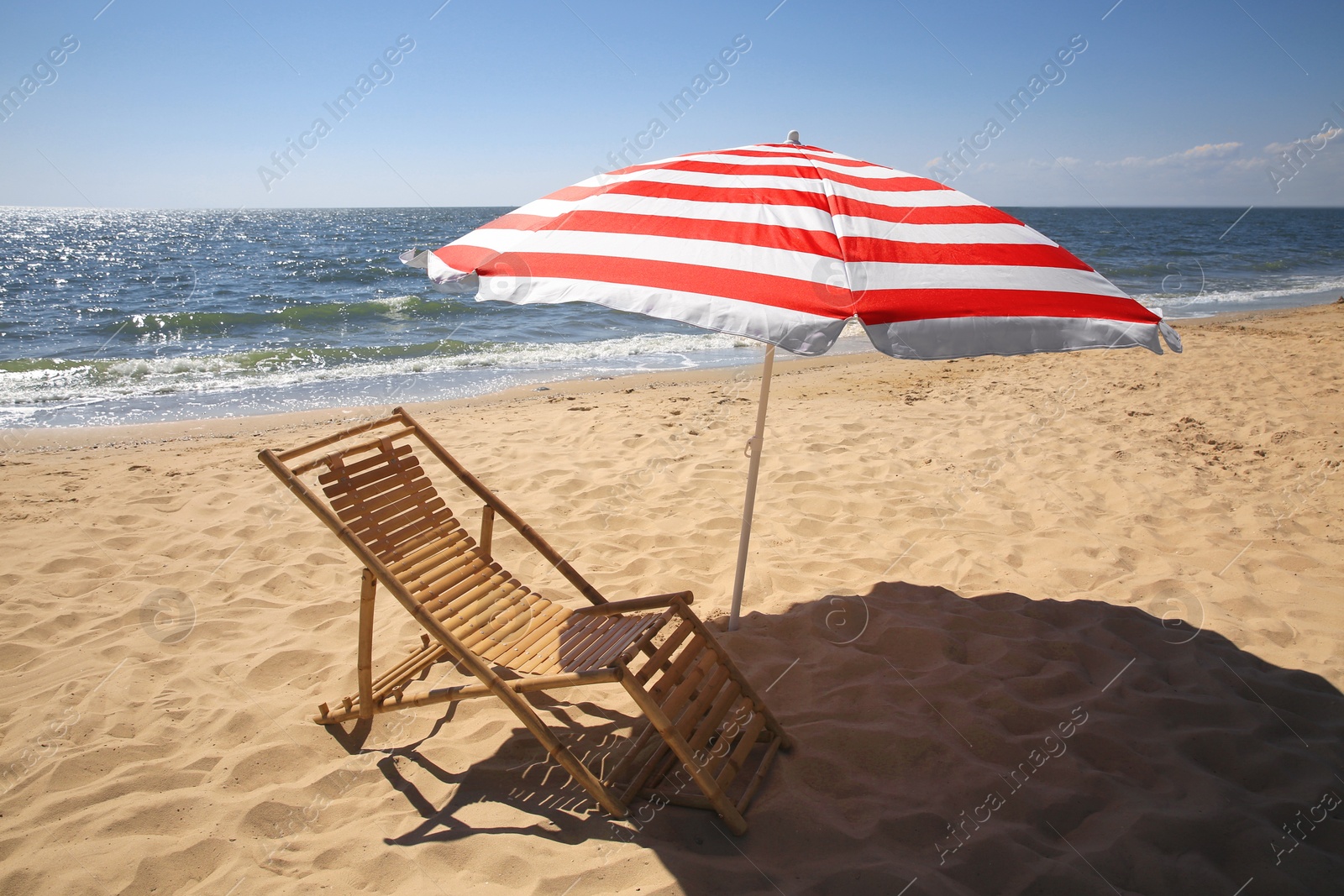 Photo of Deck chair near red and white striped beach umbrella on sandy seashore