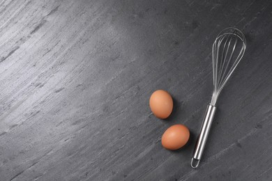 Photo of Metal whisk and raw eggs on dark grey table, flat lay. Space for text