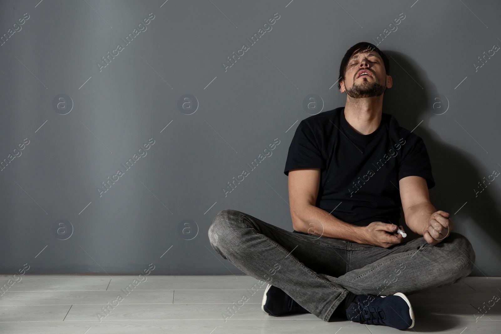 Photo of Male drug addict with syringe sitting near grey wall. Space for text