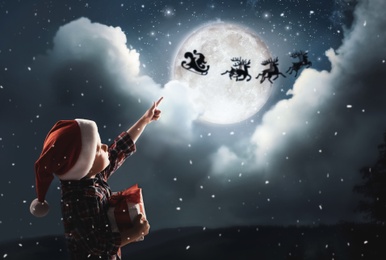 Image of Cute little boy looking at Santa Claus with reindeers in sky on full moon night. Christmas holiday