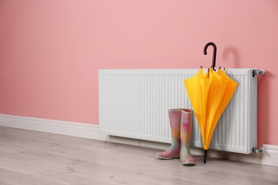 Photo of Modern radiator, rubber boots and umbrella near color wall with space for text. Central heating system