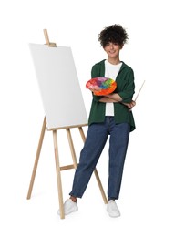 Photo of Young woman holding brush and artist`s palette near easel with canvas against white background