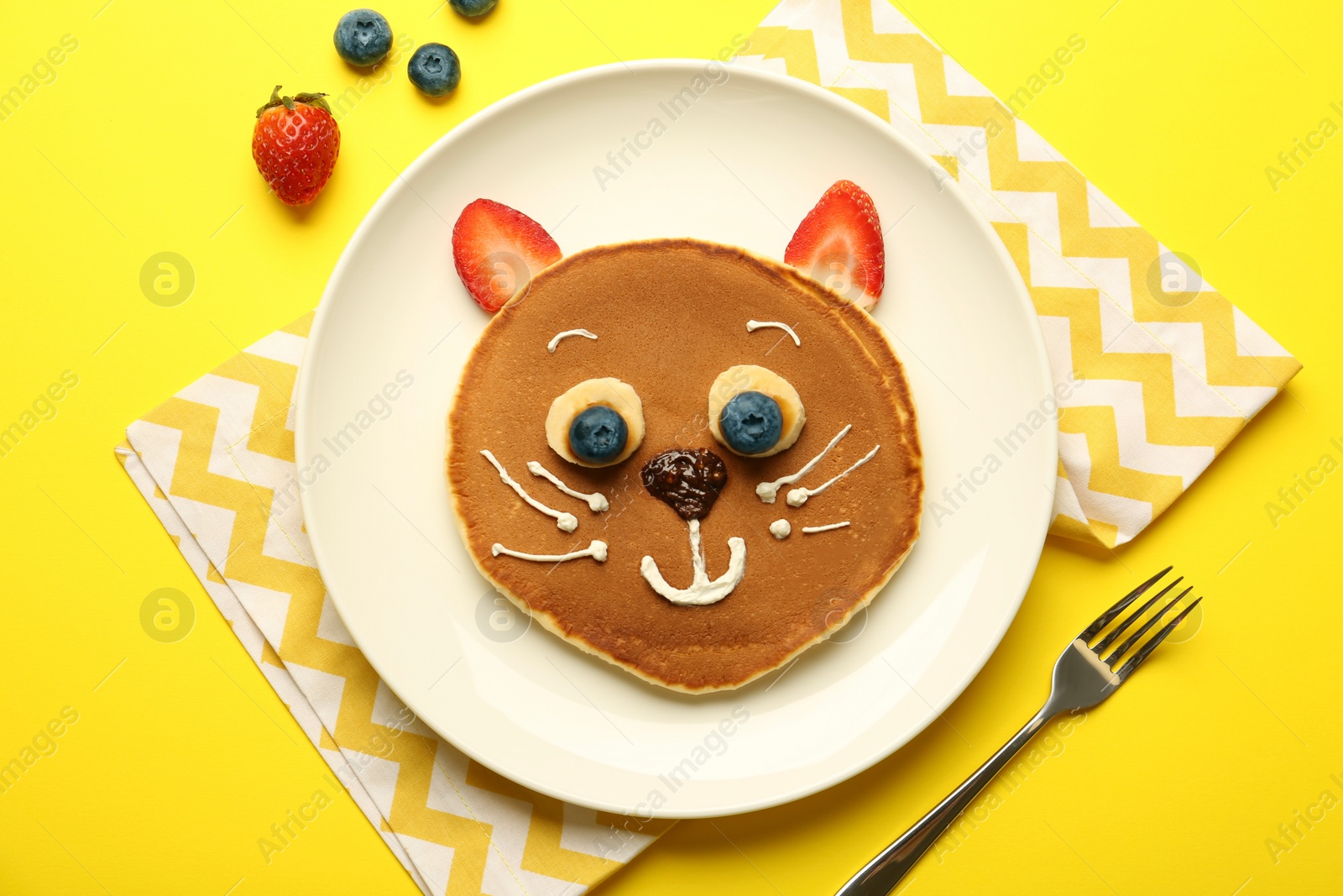 Photo of Creative serving for kids. Plate with cute cat made of pancakes, berries, cream, banana and chocolate paste on yellow background, flat lay