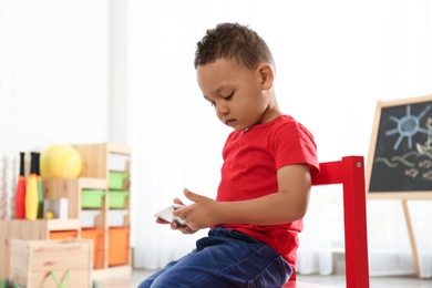 Photo of Cute little African-American child playing on mobile phone in kindergarten, space for text. Indoor activity