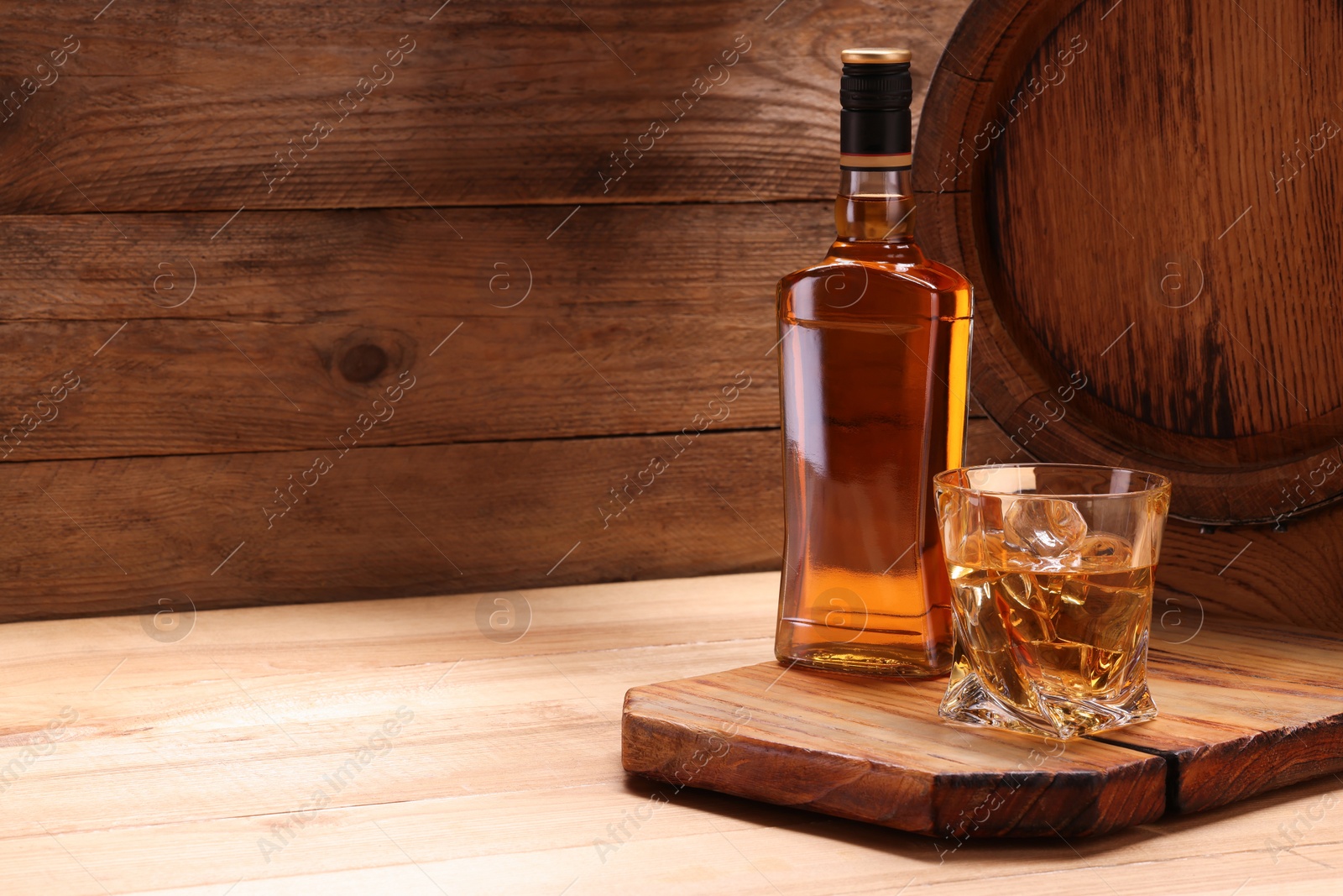 Photo of Whiskey with ice cubes in glass, bottle and barrel on wooden table, space for text