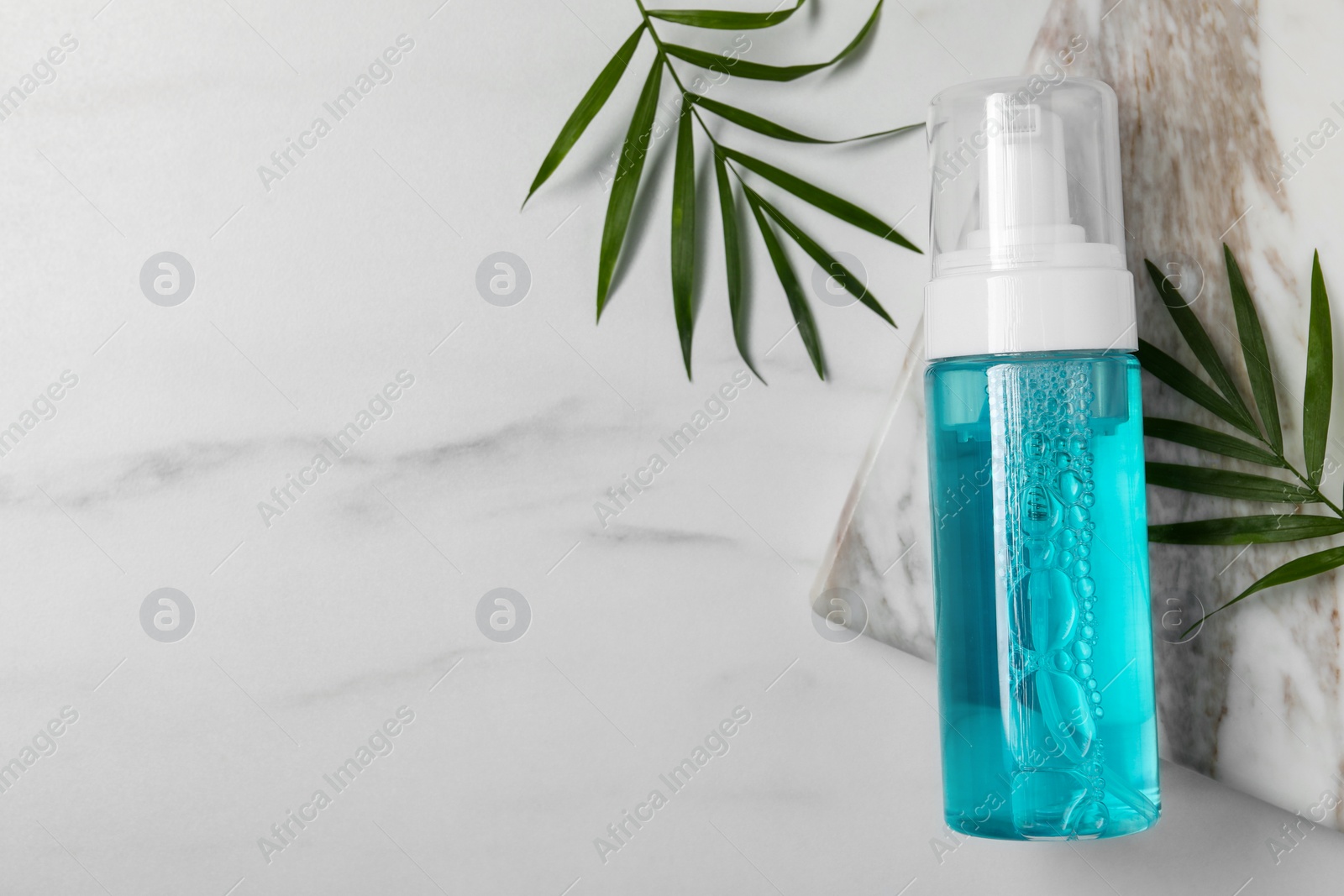 Photo of Bottle of face cleansing product and green leaves on white marble table, flat lay. Space for text
