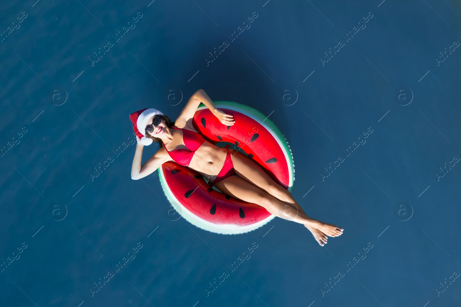 Image of Young woman wearing Santa hat and bikini with inflatable ring in sea, top view. Christmas vacation