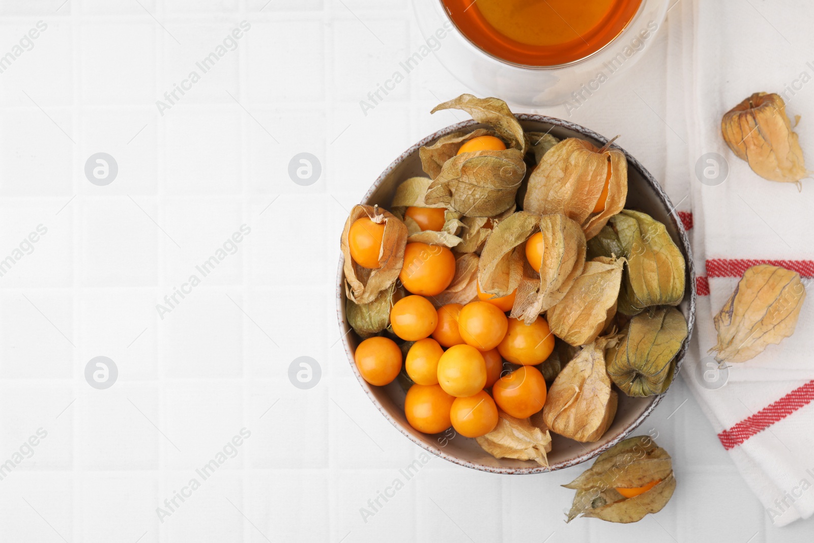 Photo of Ripe physalis fruits with calyxes in bowl on white tiled table, top view. Space for text